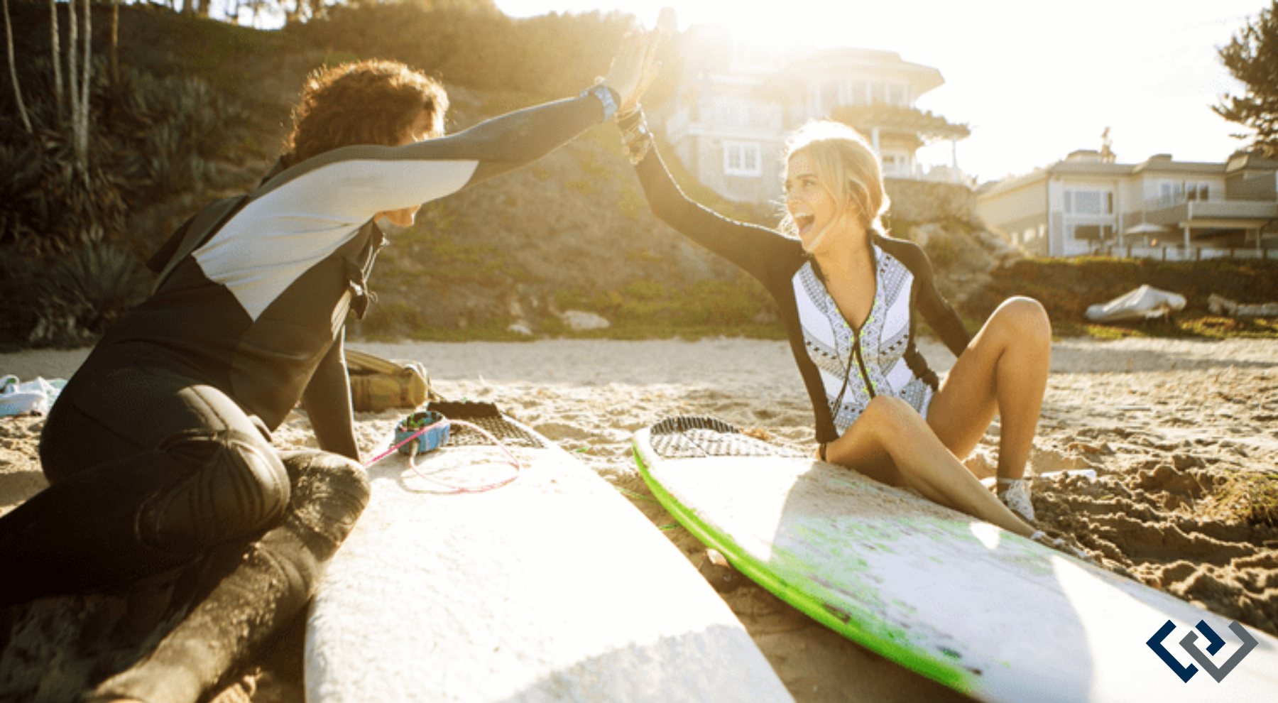 two people on the beach sitting on surfboards, giving a high-five