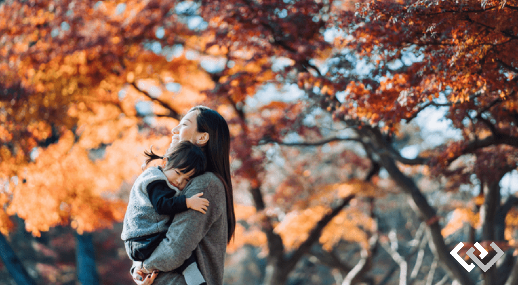 A woman holding a young child with a background of trees trimmed with autumn leaves